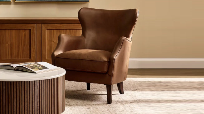 Close-up of a classic brown leather wingback armchair next to a round, slatted wood and marble coffee table