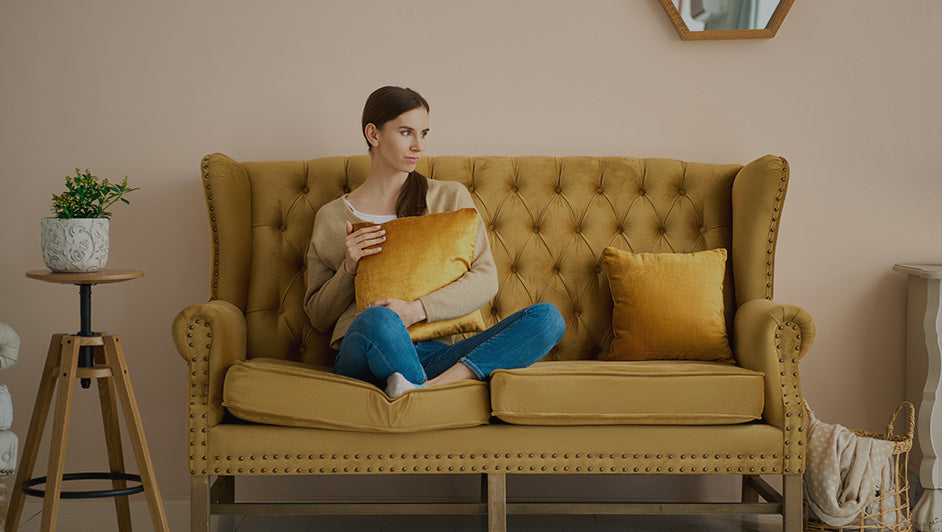 Woman sitting on a mustard yellow tufted sofa holding a matching velvet cushion, showing how cushions transform a living space.